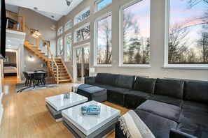 A spacious living area with a large black sectional sofa, a coffee table, and a dining area with a table and chairs. A wooden staircase leads to an upper level. - The Fairchild Haus | 30 Acres, Sauna, EV Charger (Logan)