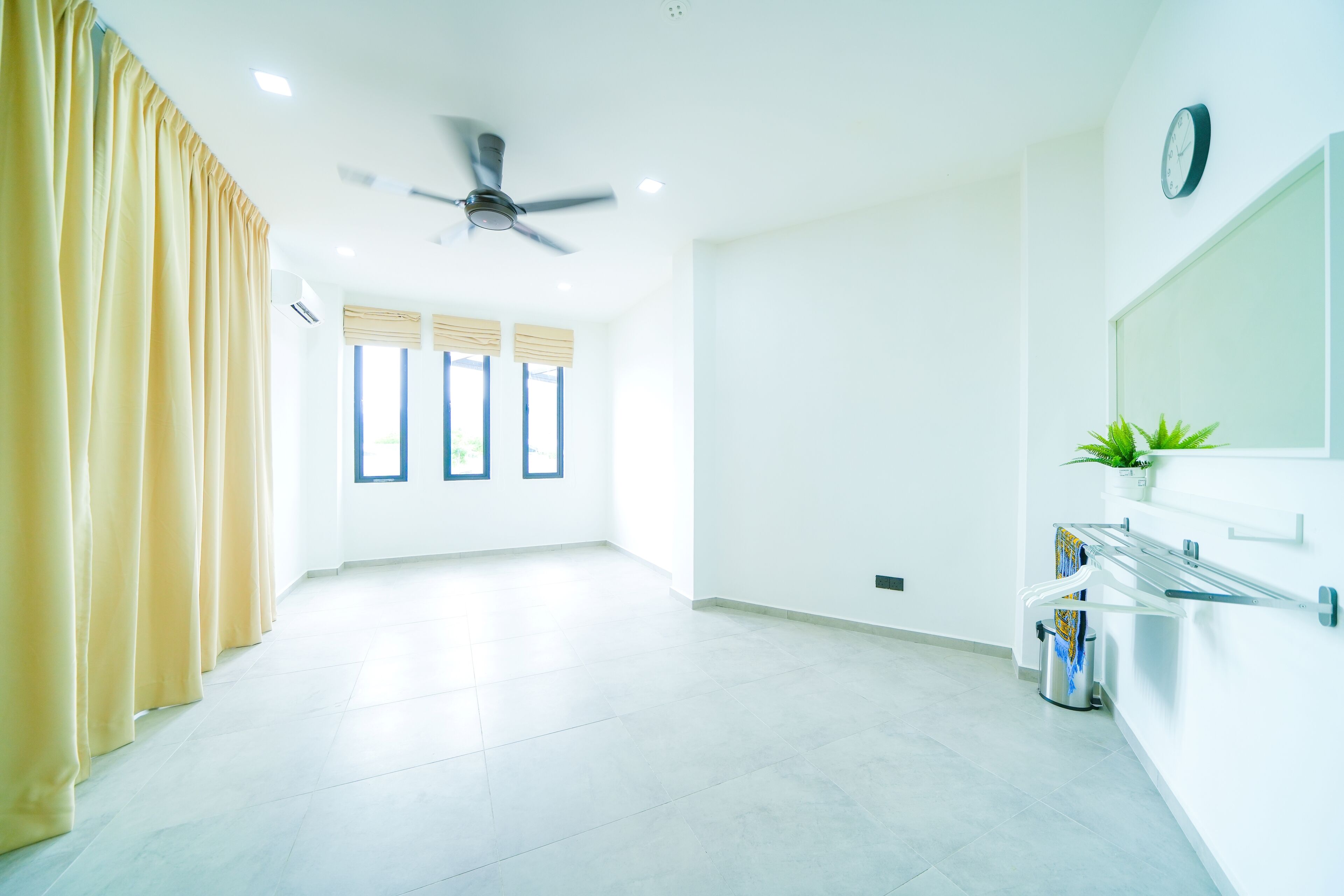 A bright, spacious room with a ceiling fan, large windows with blinds, a potted plant, and a wall-mounted mirror.