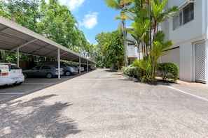 A covered parking area with several cars parked, surrounded by lush greenery and a building with a white exterior and lattice window design.