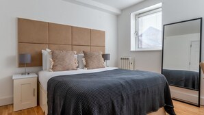 A modern bedroom with a large bed, a headboard, bedside lamps, a nightstand, a radiator, a window with blinds, and a mirror. - Divine Canary Wharf 1BR w/ Parking, nr Food & Station, by Blueground (London)
