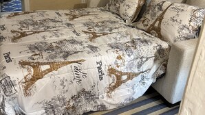 Une chambre d’hôtel avec un lit recouvert d’une couette aux motifs parisiens, un tapis rayé et une vue sur le couloir.