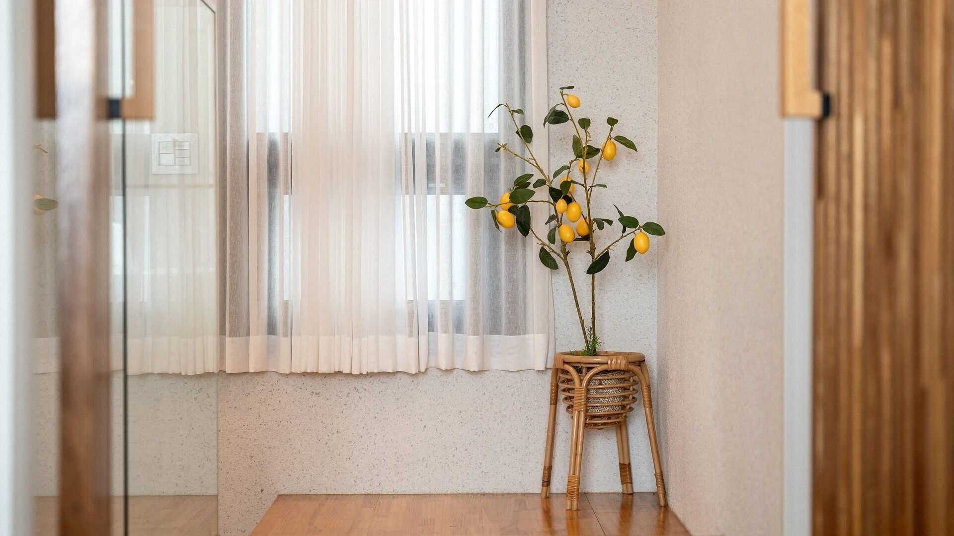 A room with a wooden table, a wicker chair, a potted plant with yellow flowers, and sheer curtains.
