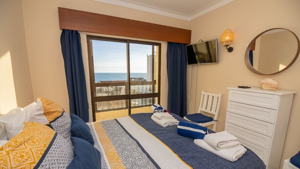 A hotel room with a large bed, a dresser, a TV, and a balcony with a view of the sea. - Apartamento Mar Azul (Portimão)