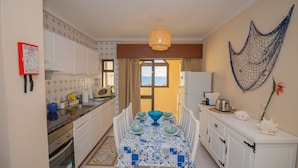 A compact kitchen with white cabinets, a dining table with blue and white tablecloth, and a view of the sea through the window. - Apartamento Mar Azul (Portimão)