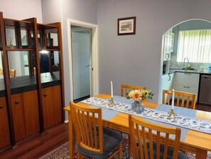 A dining area with a wooden table, chairs, a vase with flowers, a candle, and a kitchen in the background.