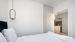 Une chambre d’hôtel moderne avec un lit soigneusement fait, une suspension lumineuse et une vue sur la cuisine.