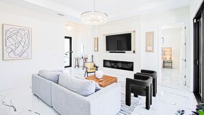 A modern living room with a grey sofa, a wooden coffee table, and a TV mounted on the wall. There is a fireplace below the TV and a chandelier hanging from the ceiling.