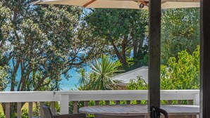 A wooden deck with wicker furniture and a white railing, overlooking a lush garden and a body of water. - Miro Beach House at Palm Beach (Waiheke Island)