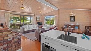 A spacious living area with a fireplace, a dining area, and a kitchen with a large sink. - Miro Beach House at Palm Beach (Waiheke Island)