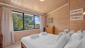 A wooden-paneled bedroom with a large bed, a dresser, and a window with curtains. - Miro Beach House at Palm Beach (Waiheke Island)
