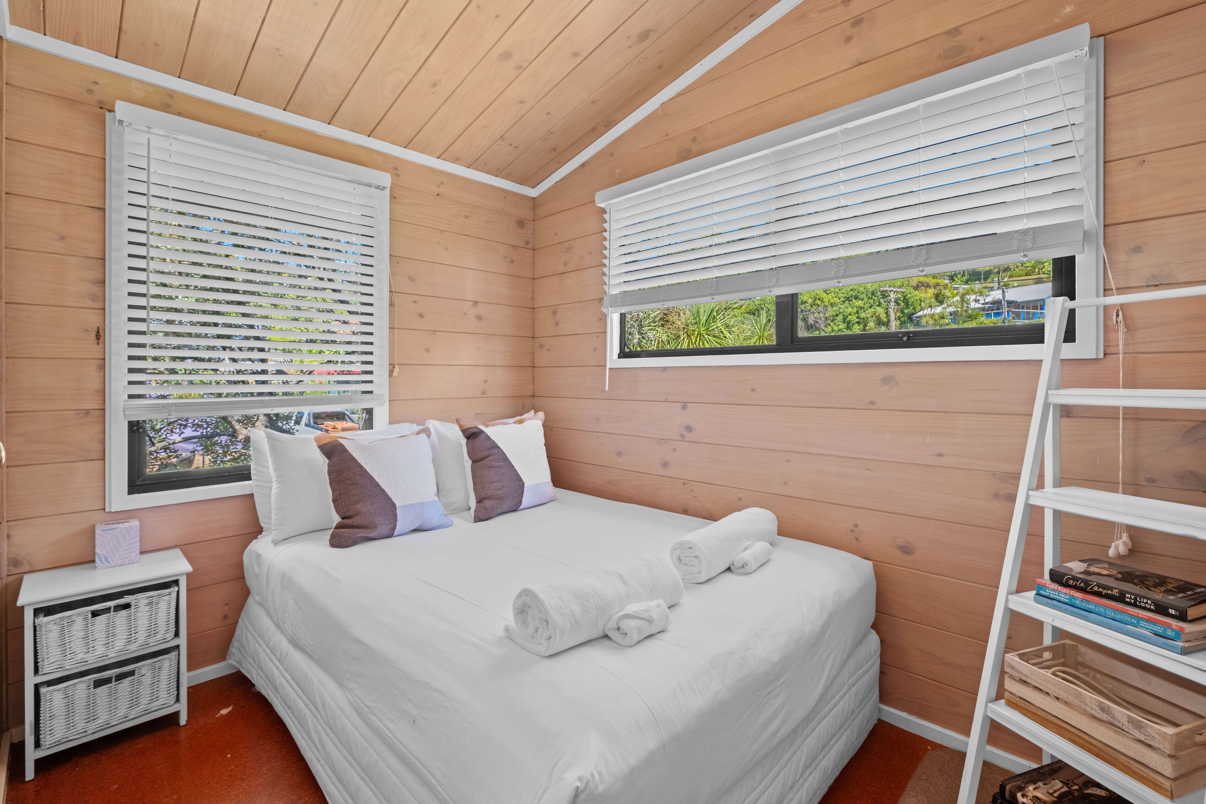 A bedroom with wooden walls, a bed with white bedding, two windows with blinds, a bedside table with a lamp, and a small shelf with books.