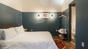 Un dormitorio con una cama, un aplique en la pared, un perchero y una puerta que da a un baño.