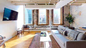 A modern living room with a sofa, a coffee table, and a TV. There are three windows with views of buildings. - Spacious Loft, 2 Blocks from Broadway! (Loft 302) (Nashville)