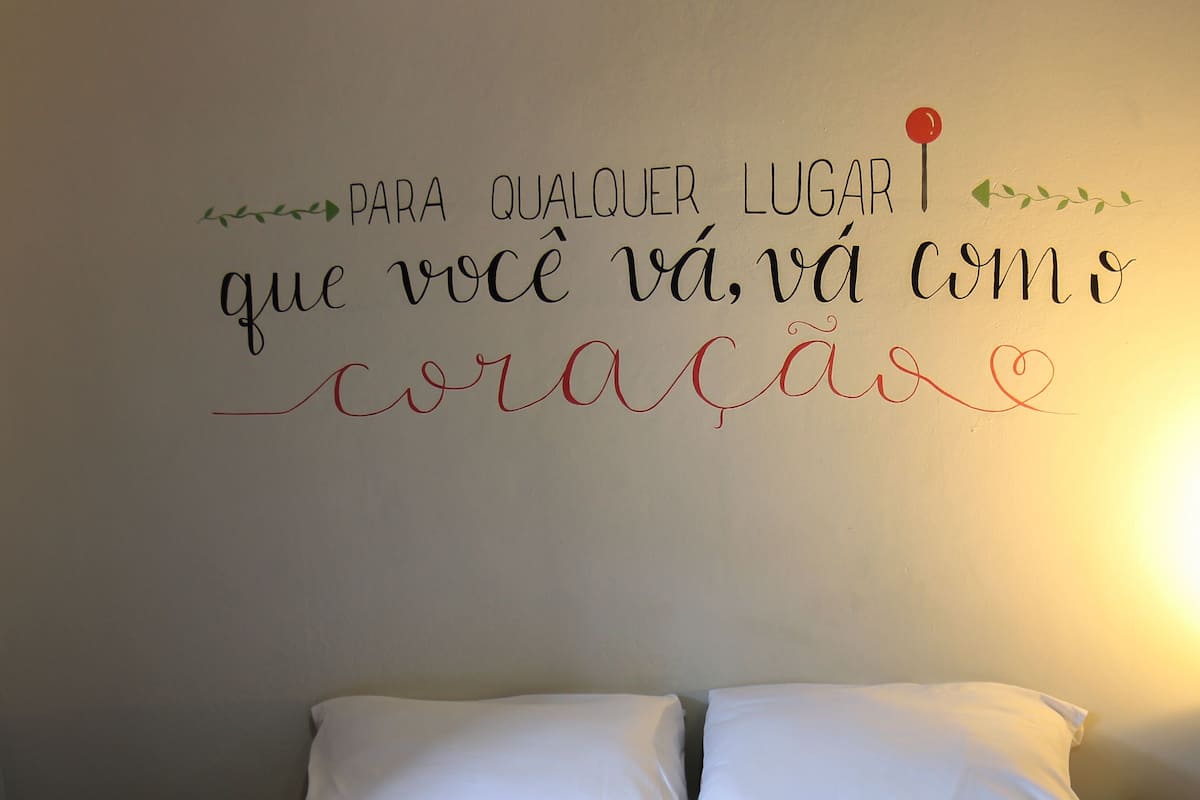 A neatly made bed with white linens and rolled towels, a wall with a motivational quote in Portuguese, and a simple, clean room design.