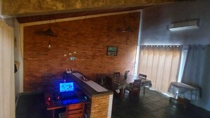 A room with a brick wall, a wooden desk with a computer, a dining table with chairs, and a hanging light fixture.