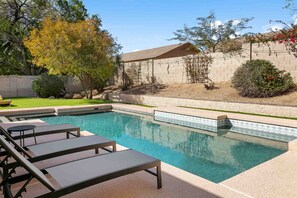 A rectangular swimming pool with a tiled edge, surrounded by a beige wall and a small garden area with a few trees and shrubs. - SWSS Upscale Kierland 4BR Retreat | Heated Pool, Outdoor Living, Gourmet Kitchen & Fire Pit (Scottsdale)