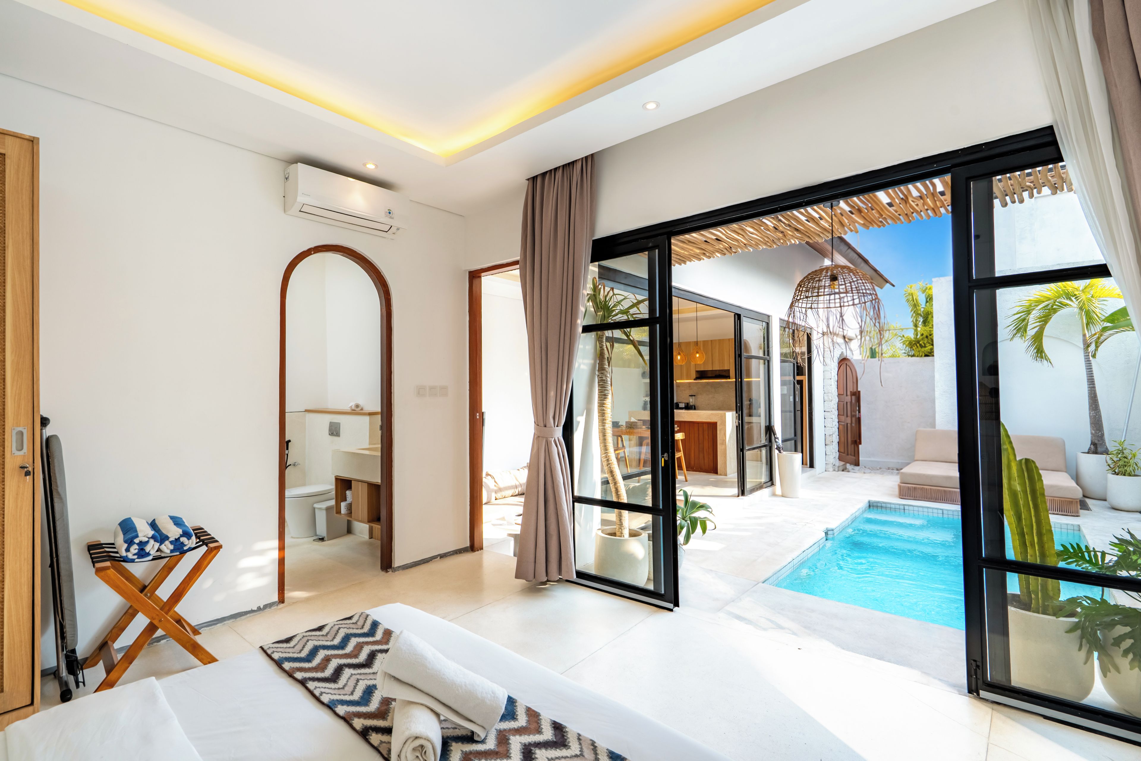 A modern hotel room with a large sliding glass door leading to a pool area, a separate dining area, and a bathroom with a mirror and towel rack.