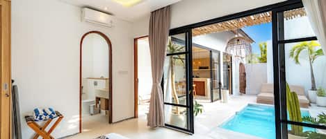 A modern hotel room with a large sliding glass door leading to a pool area, a separate dining area, and a bathroom with a mirror and towel rack.
