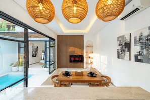 A modern living room with a dining area, a TV mounted on a wooden wall, and a pool visible through the glass door. - Yahvi Villa by Betterplace (Canggu)