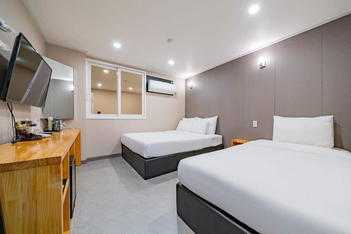 A hotel room with two beds, a wooden desk, a TV, and an air conditioning unit.