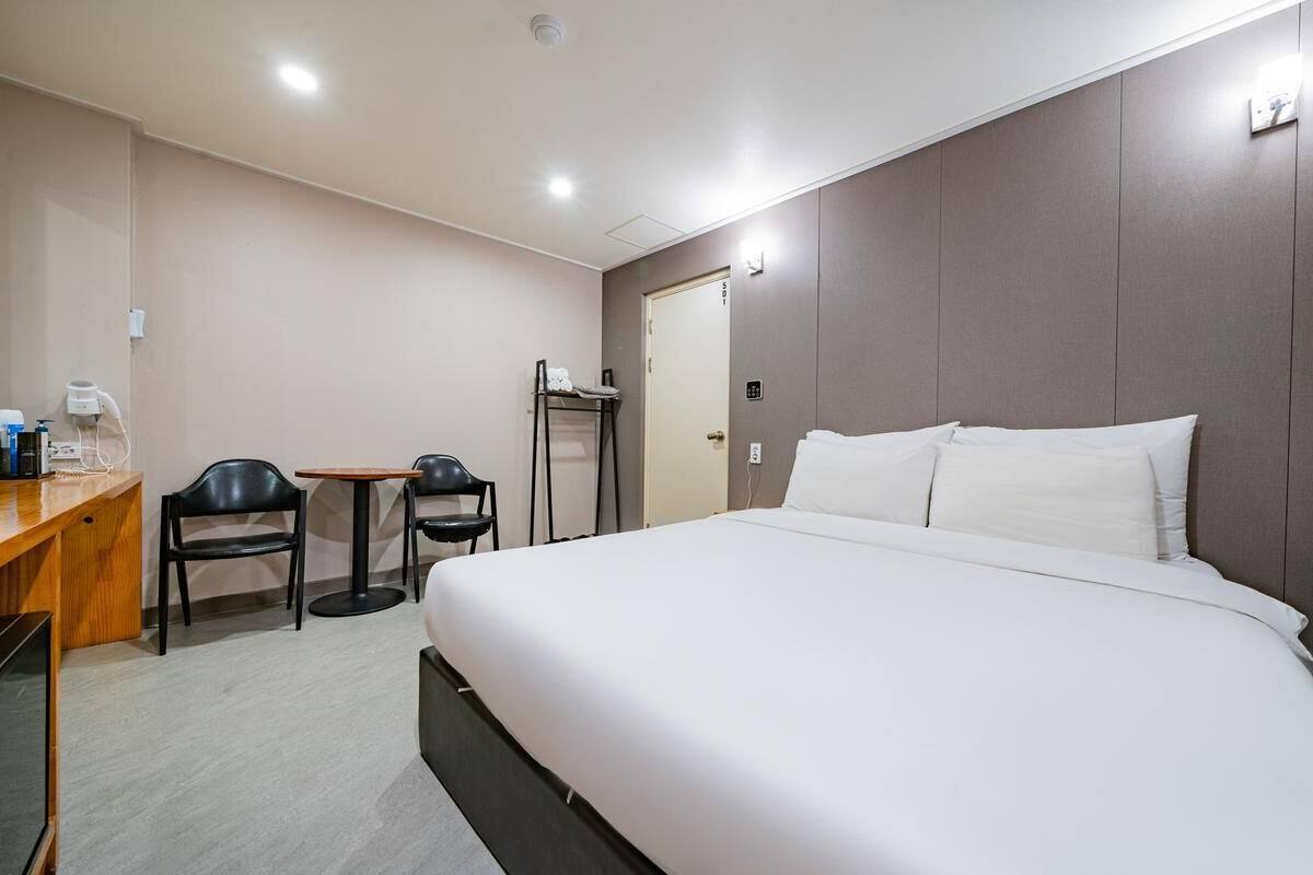 A hotel room with a large bed, a desk with a chair, a small table, and a wardrobe.