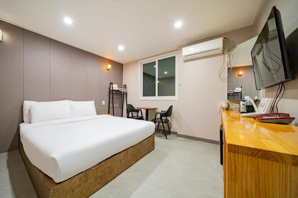 A hotel room with a bed, a desk, a chair, a TV, and an air conditioning unit.