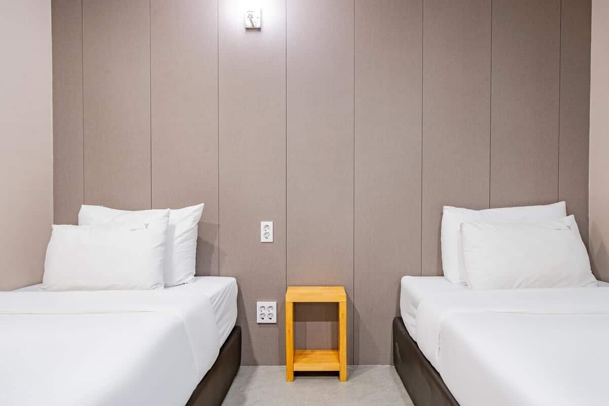 A hotel room with two beds, a small wooden nightstand, and a light switch on the wall.