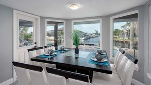A dining area with a dark wooden table, white chairs, and a view of a marina through large windows. - Gulf Access, Beautiful Water Views, Paddle Board, Heated Pool - Villa Manhattan - Roelens (Cape Coral)