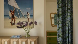 A room with a geometric wooden cabinet, a framed beach painting, a vase with purple flowers, a green dresser, and a blue curtain with a tropical pattern. - NEW Wellness Focused Adult Only Boutique Hotel - Standard Suite ADA Compliant (Rodanthe)
