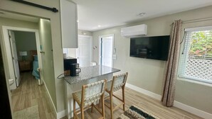 A compact kitchen with a granite countertop, two bar stools, a built-in TV, and a window with patterned curtains. - Turtle Nest 2 - Quick Walk to Beach (Fort Myers Beach)