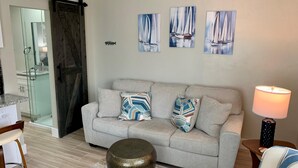 A living room with a grey sofa, colorful pillows, a round ottoman, and three paintings of sailboats on the wall. - Turtle Nest 1 - Quick Walk to Beach (Fort Myers Beach)