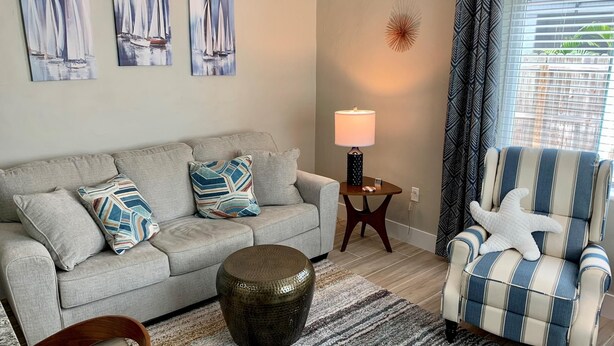 A living room with a grey sofa, striped armchair, decorative pillows, a round table with a lamp, and a starfish decoration. - Turtle Nest 1 - Quick Walk to Beach (Fort Myers Beach)