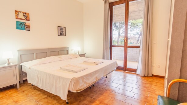 Una cama bien hecha con sábanas blancas, una mesita de noche con lámpara, y vistas al exterior a través de un ventanal.