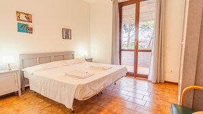 Ein ordentlich bezogenes Bett mit weißen Laken, ein Nachttisch mit Lampe und Blick ins Freie durch eine Glastür.