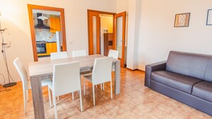 Un comedor con una mesa de madera y sillas blancas, un sofá gris, y una cocina visible a través de una puerta abierta.