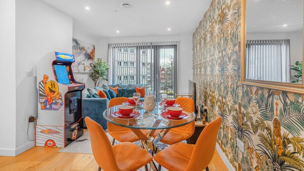 Una sala de estar moderna con una mesa de comedor de vidrio, sillas naranjas, un sofá azul y una máquina arcade retro.