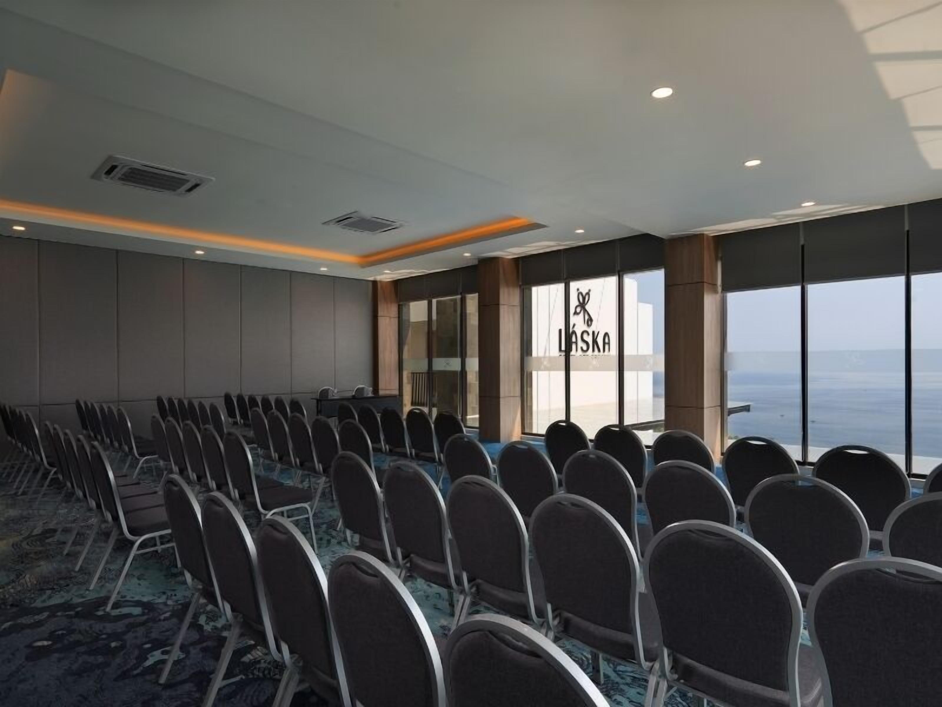 Uma sala de conferência com fileiras de cadeiras, janelas amplas com vista e um logotipo na parede.