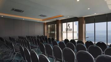 Uma sala de conferência com fileiras de cadeiras, janelas amplas com vista e um logotipo na parede.