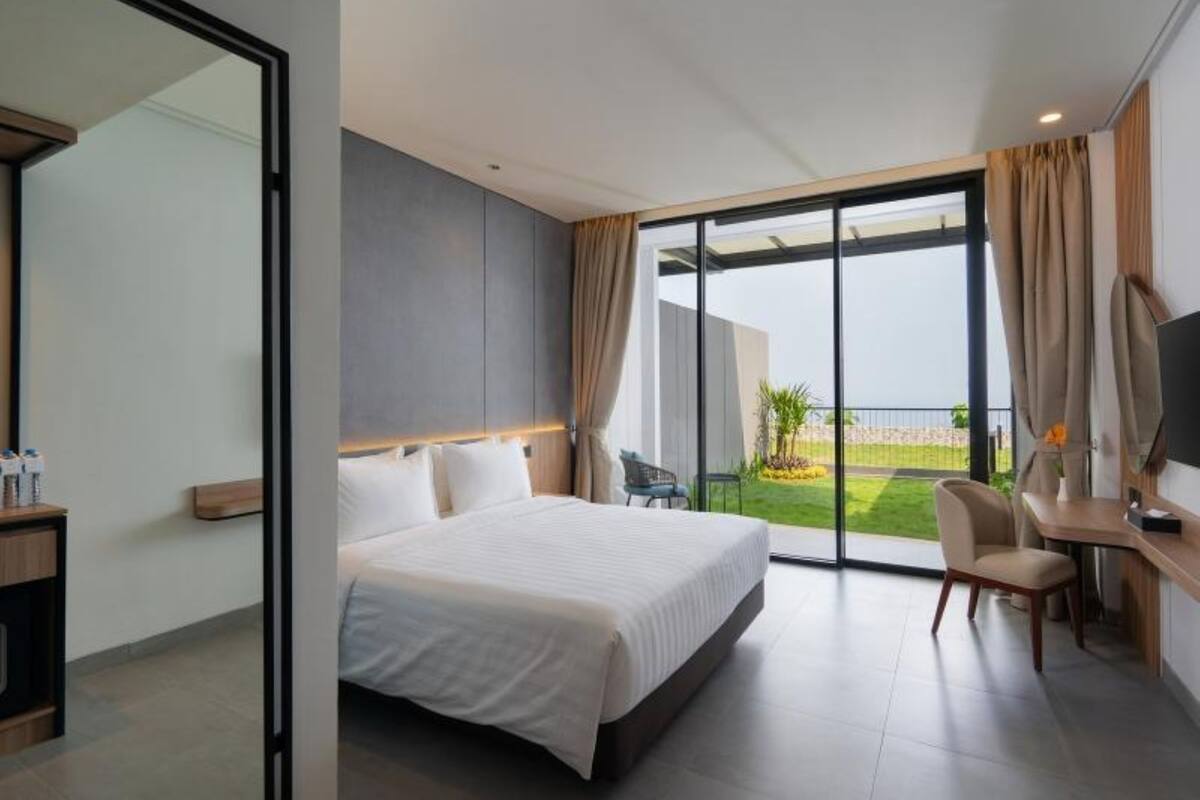 A modern hotel room with a large bed, a desk, and a TV. The room has a large window with a view of the beach.