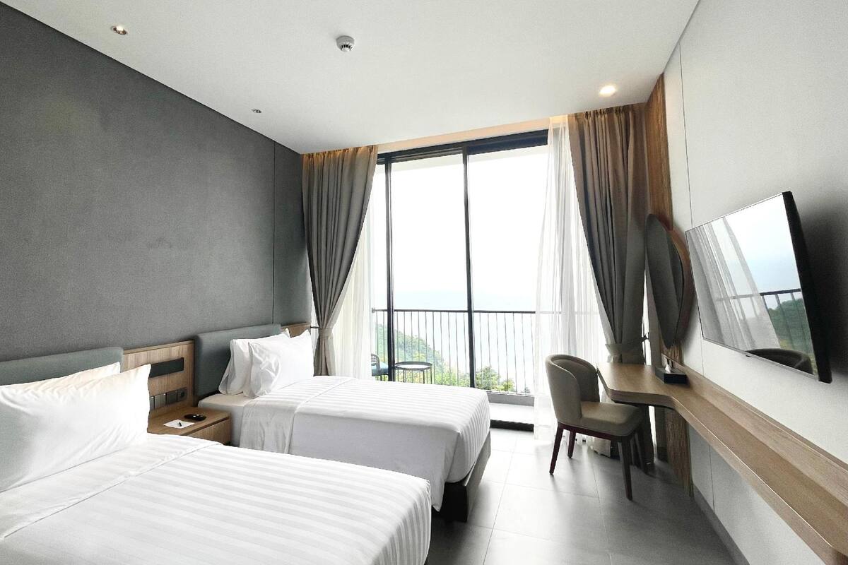 A hotel room with two beds, a desk, a chair, a TV, and a balcony with a view.