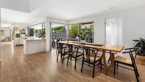 A modern kitchen with a central island, dining area with wooden chairs, and a view of the garden through large windows. - Luxury coastal retreat only 250m from the beach! (Tweed)