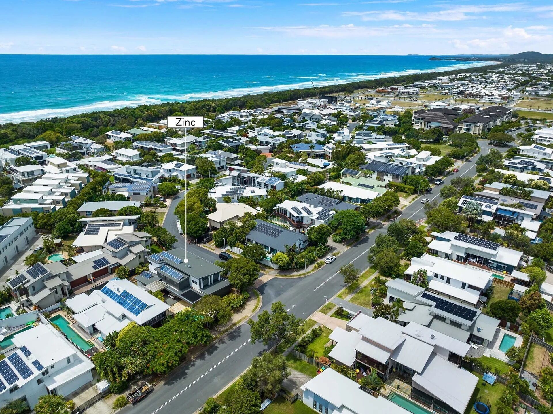 A residential area with houses featuring solar panels, a road, and a beach in the background.