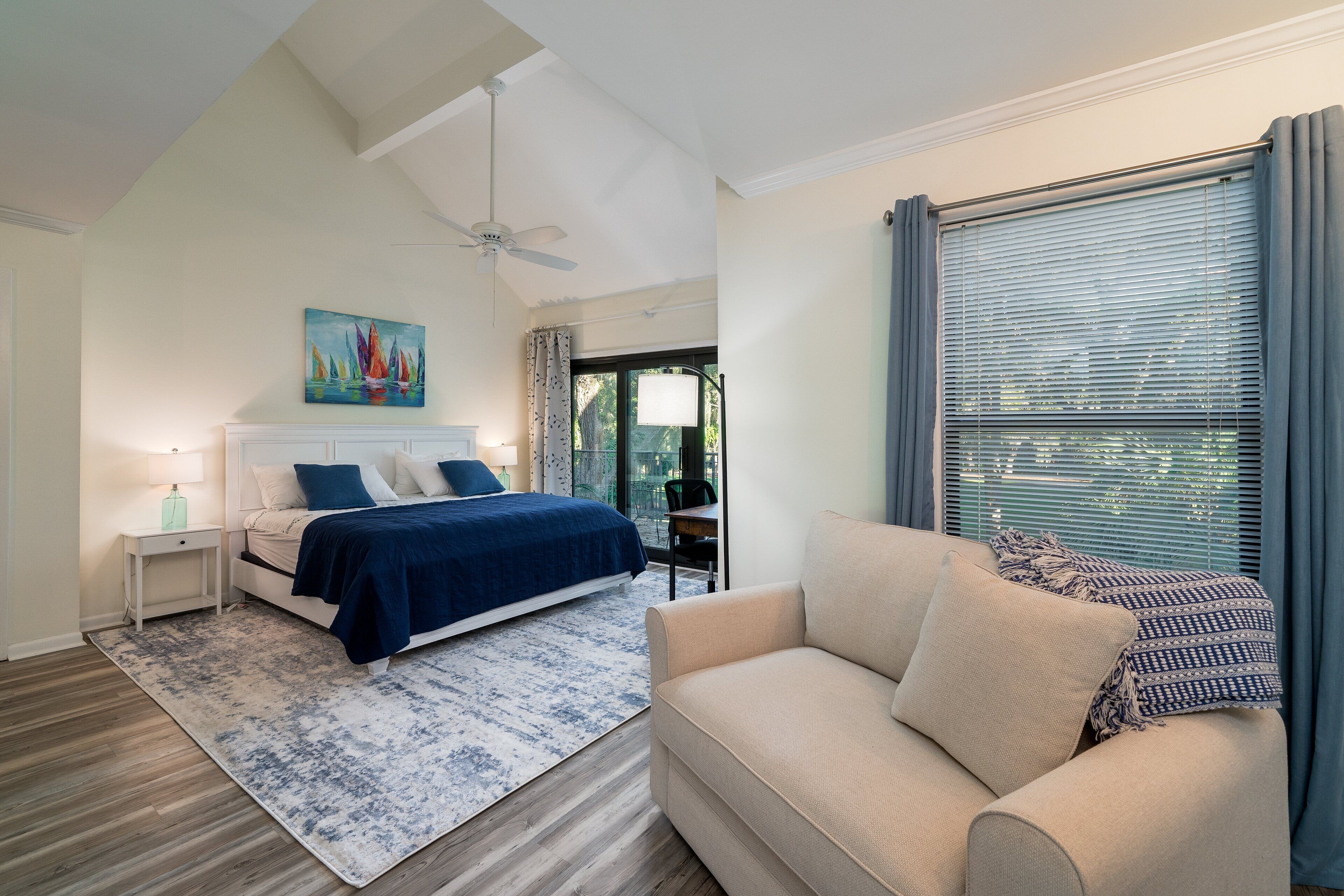 Une chambre à coucher comprenant un lit, une table de chevet, une commode, un ventilateur de plafond, une fenêtre avec des stores et une vue sur l’extérieur.