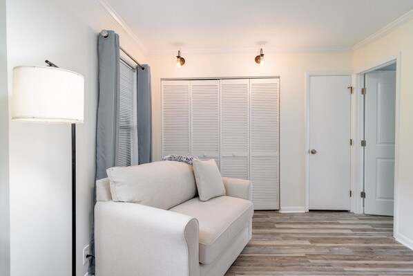 Ein neutral gestaltetes Wohnzimmer mit einer Couch, einer Stehlampe, Schränken mit Klappläden und einer Tür.