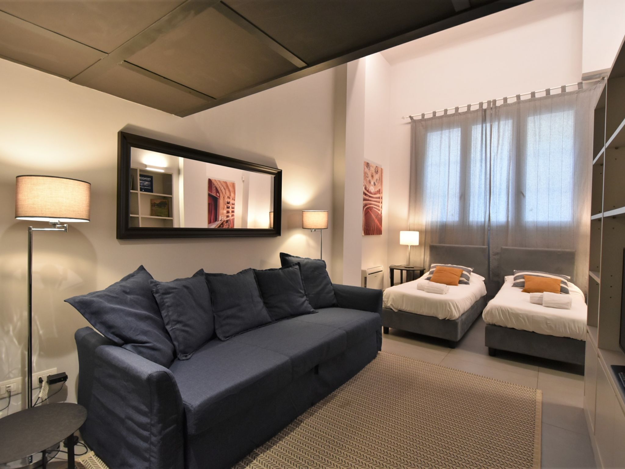 Ein modernes Hotelzimmer mit einem dunkelblauen Sofa, zwei Einzelbetten, einem großen Spiegel und Blick auf die Stadt durch transparente Vorhänge.