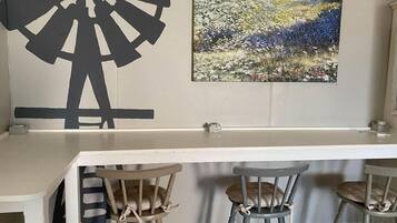 Uma área de bar com bancada branca, três banquetas, uma grande decoração de parede abstrata e uma pintura de paisagem.