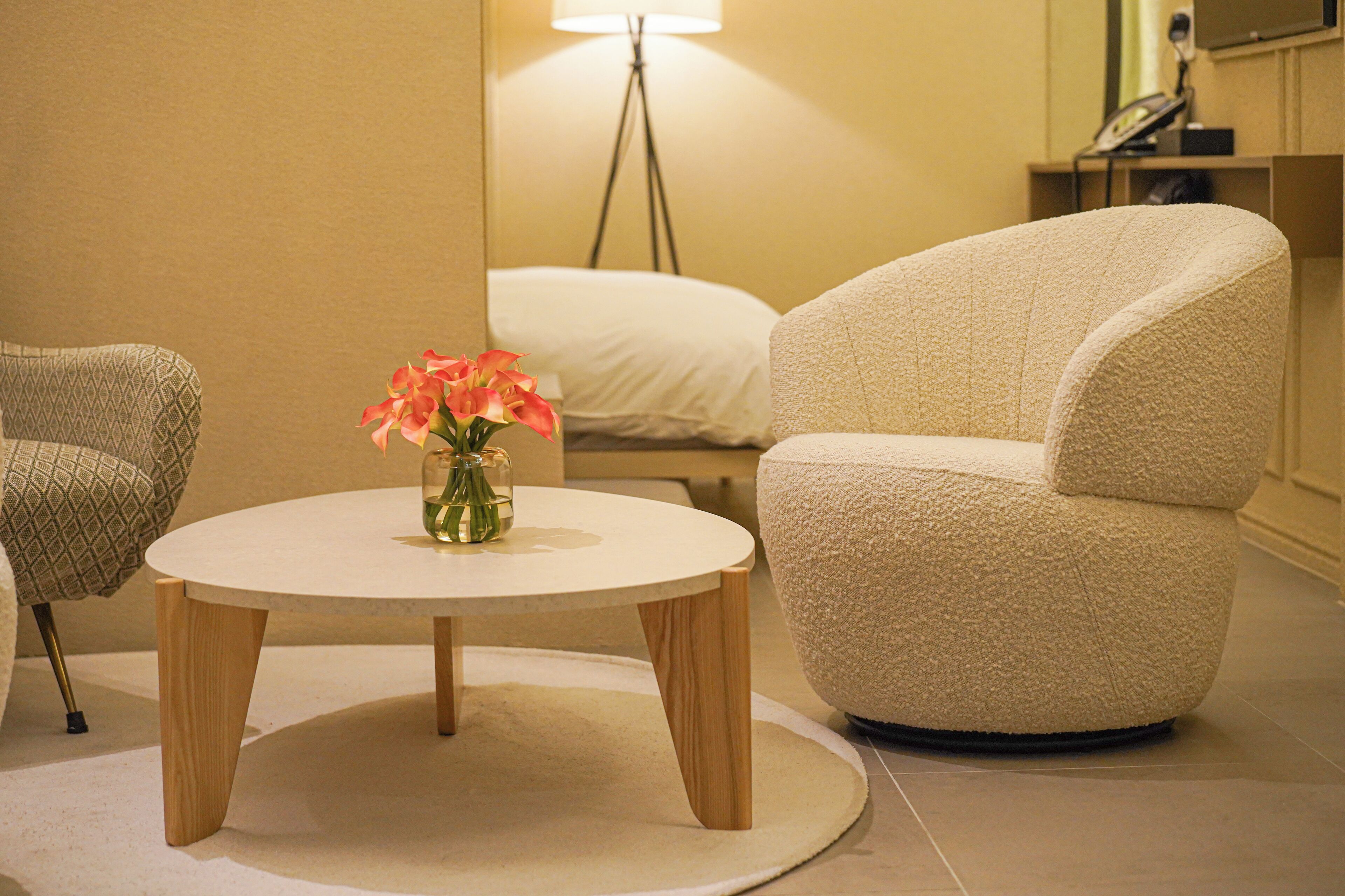 Una habitación con una mesa redonda, dos sillones, un jarrón con flores, una lámpara de pie y una cama con ropa de cama blanca.