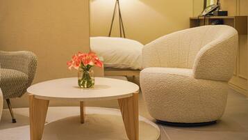 Una habitación con una mesa redonda, dos sillones, un jarrón con flores, una lámpara de pie y una cama con ropa de cama blanca.