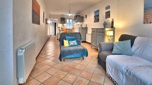 Ein geräumiges Wohnzimmer mit einer Couch, einem Sessel und einem Esstisch. Im Hintergrund ist eine Küche mit Schränken und einer Mikrowelle zu sehen.