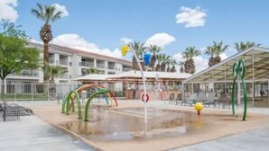 Ein Wasserspielbereich mit bunten Elementen, einem Pool und einem Gebäude mit überdachter Sitzgelegenheit im Hintergrund.
